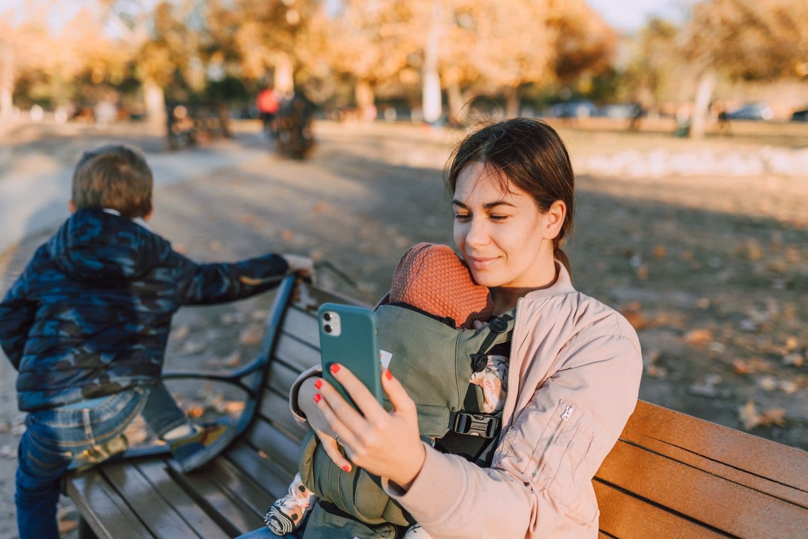A mother on a park bench with a baby secured in a structured carrier, one hand resting on the front buckle