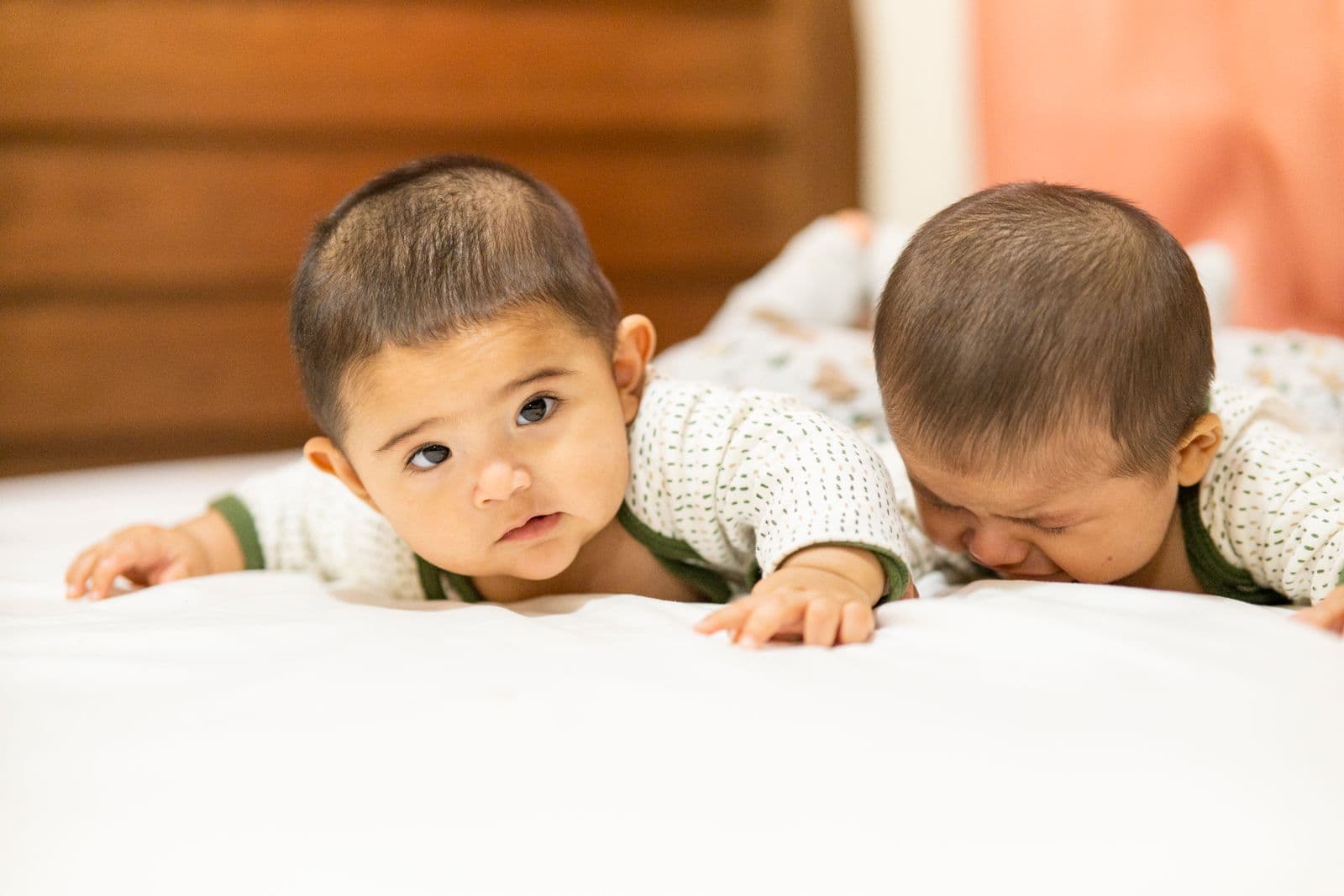 Twin babies lying side by side on a bed, one looking toward the camera