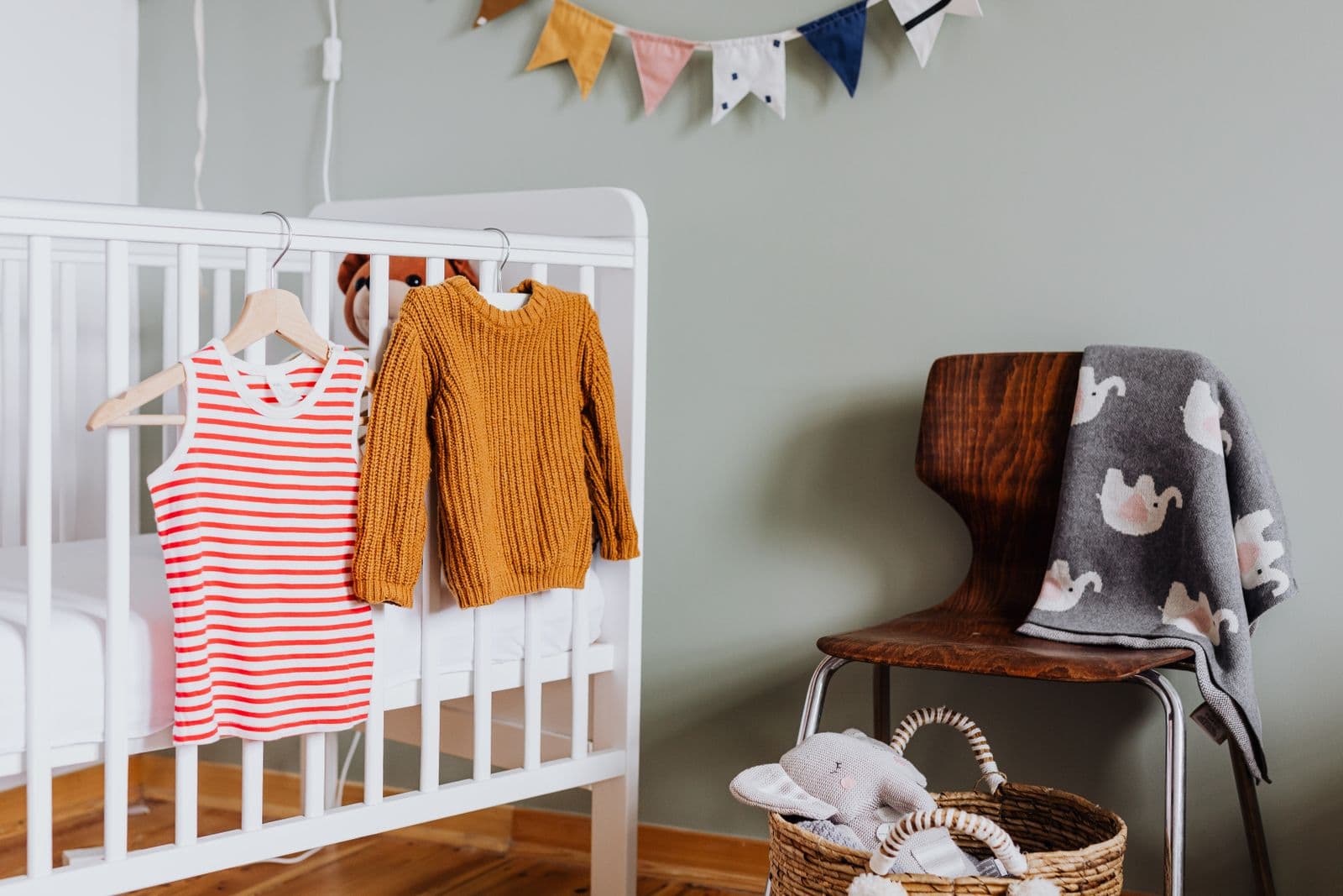 A white crib with a bare mattress and baby clothes hanging from the rail in a sage-walled nursery