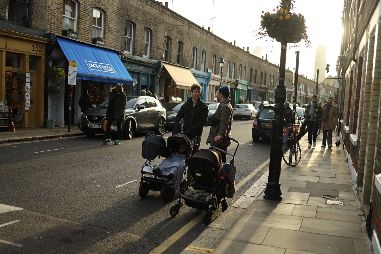 Two parents stand talking on a city street beside a side-by-side double stroller and a single stroller