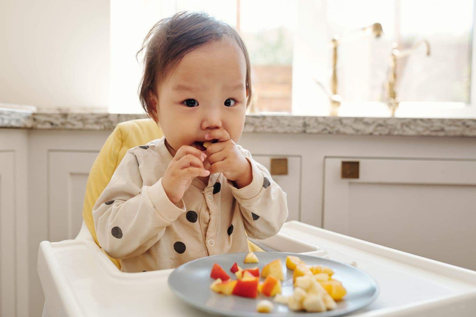 A baby in a high chair picking up pieces of cut fruit from a plate on the tray