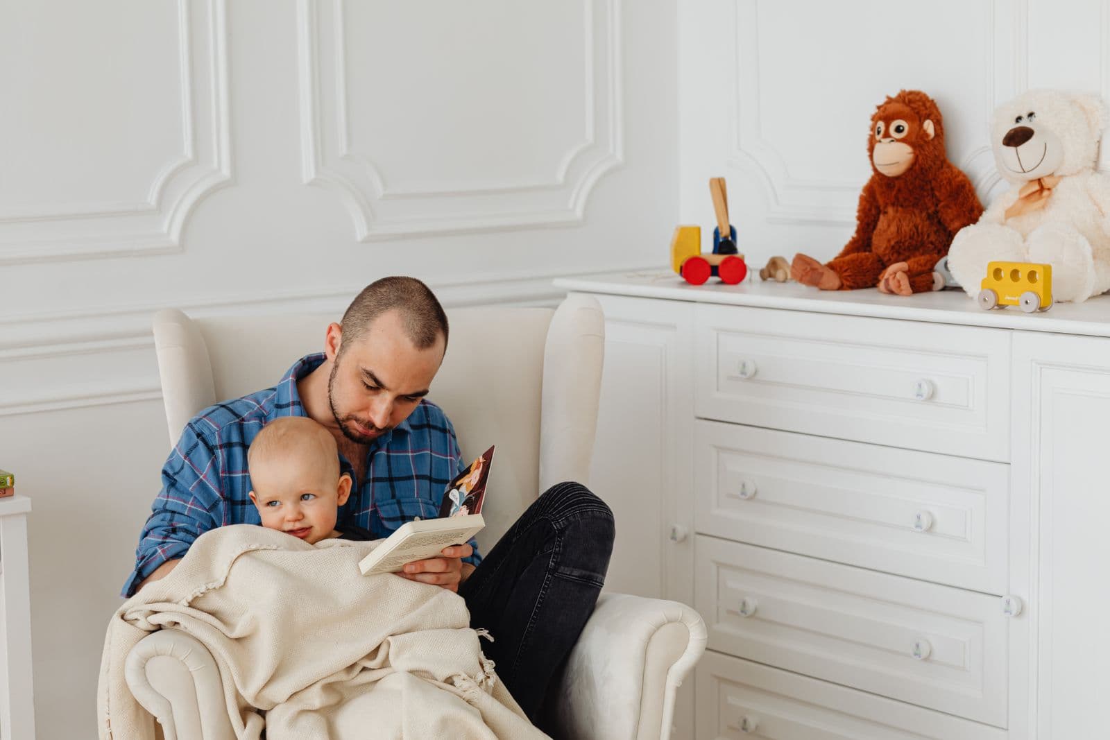 A parent sitting in an upholstered wingback nursery chair reading to a baby, with a dresser and toys behind them