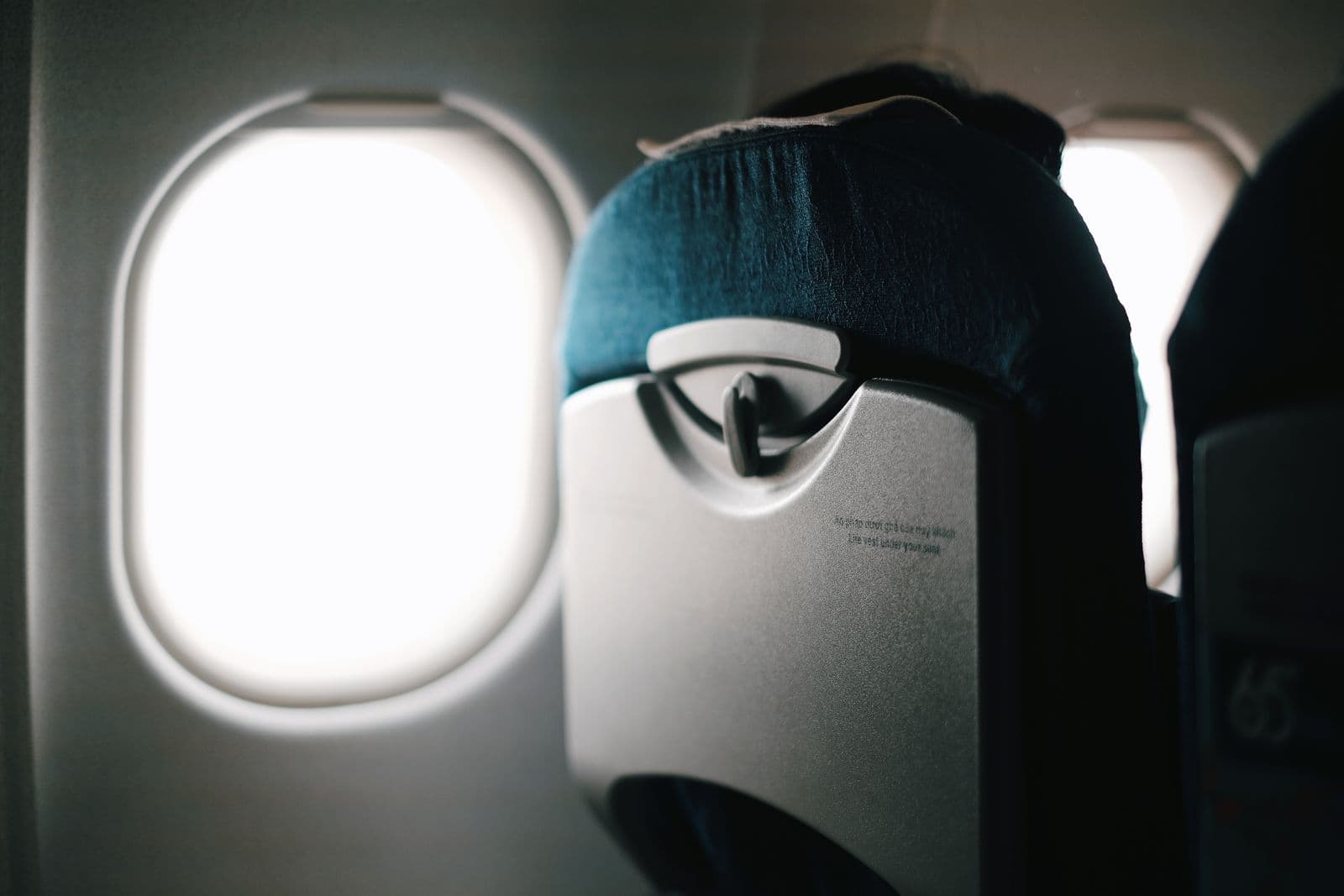 The back of an airline window seat with its stowed tray table, beside a bright cabin window