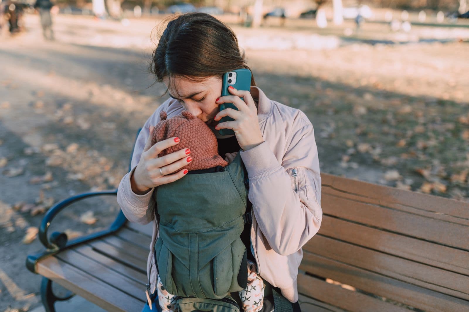 A mother on a park bench kissing the top of her baby's head, the baby facing in toward her chest in a green soft structured carrier