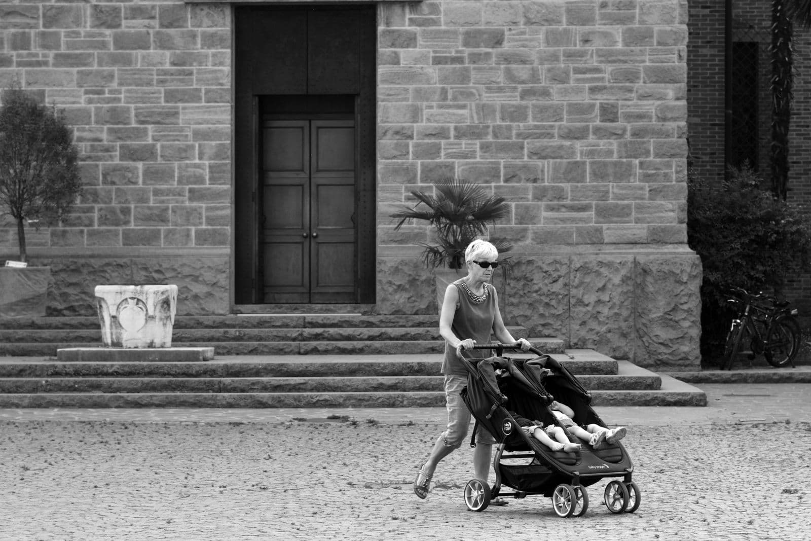 A woman pushes a side-by-side double stroller carrying two children past a stone doorway