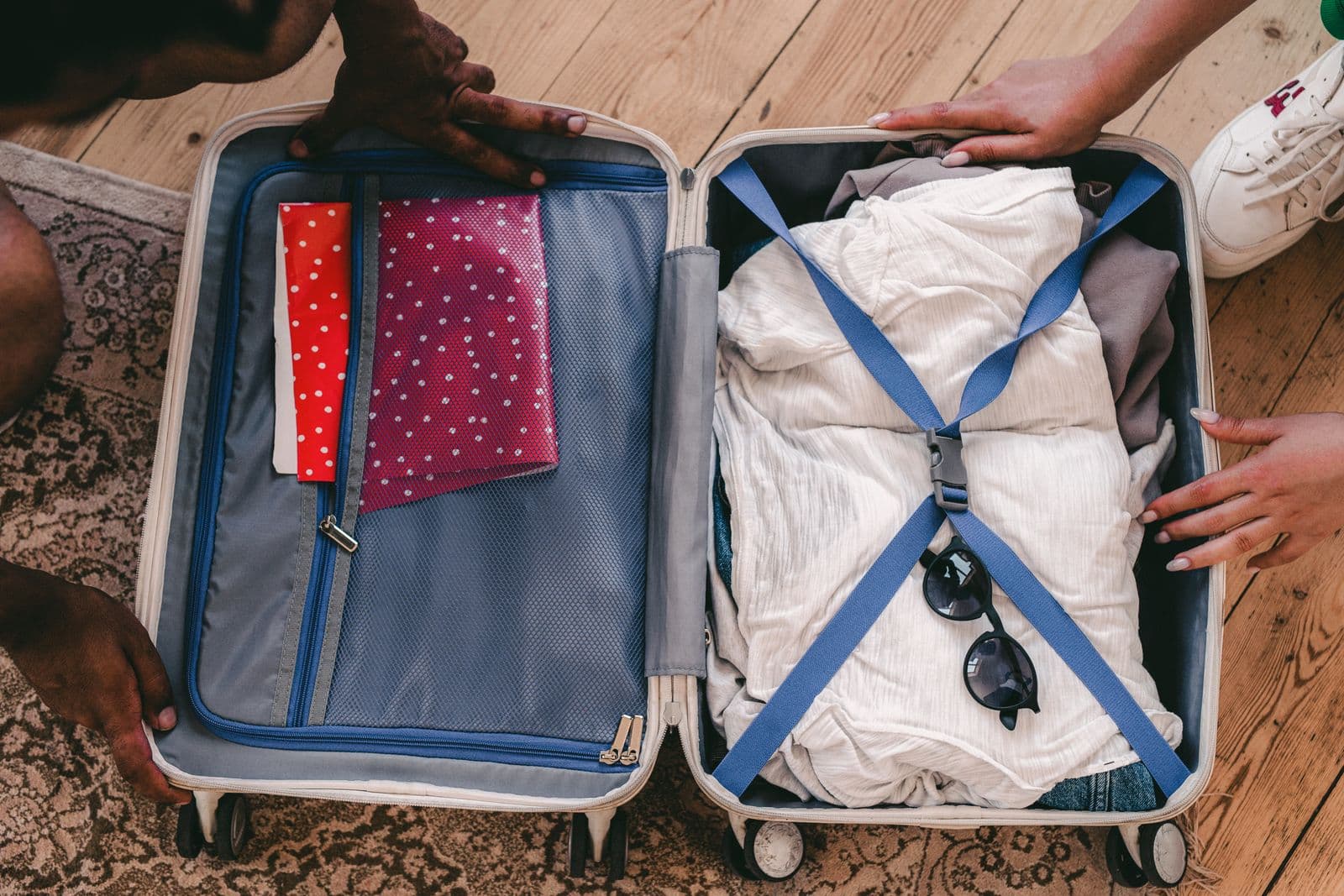 An open suitcase being packed with folded clothes and sunglasses on a wooden floor