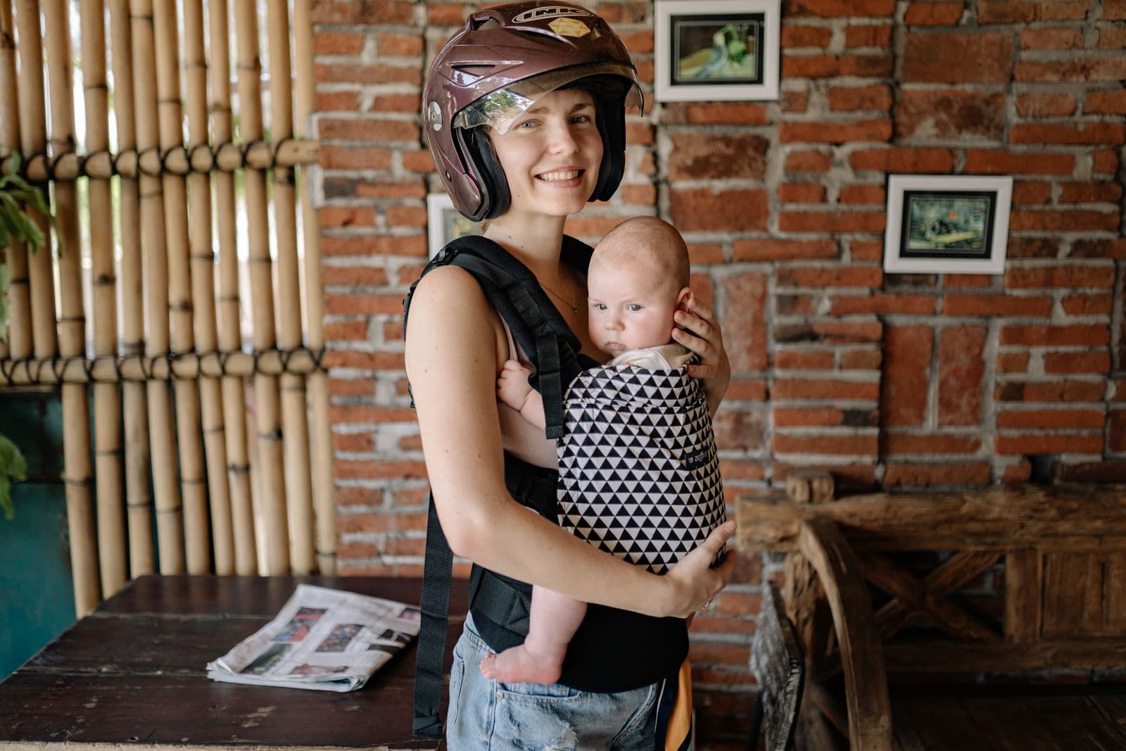 A smiling woman wearing a black soft structured carrier with her baby facing inward against her chest, the baby's face clear of the panel