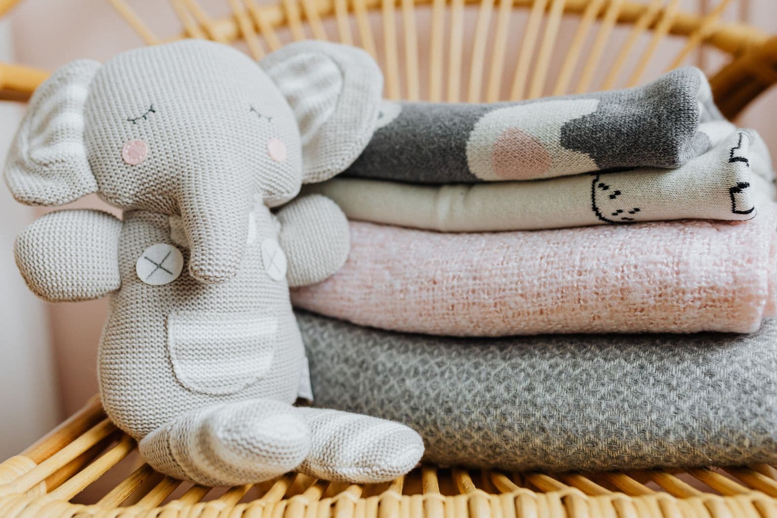 A grey knitted elephant soft toy sitting beside a stack of folded grey, cream and pink baby blankets on a wicker chair