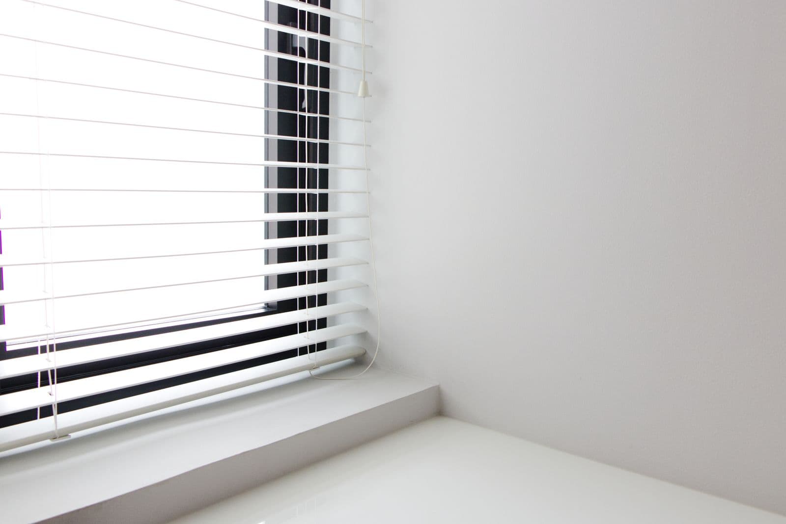 A white slatted window blind with its pull cord and cord-stop toggle hanging down beside a window ledge
