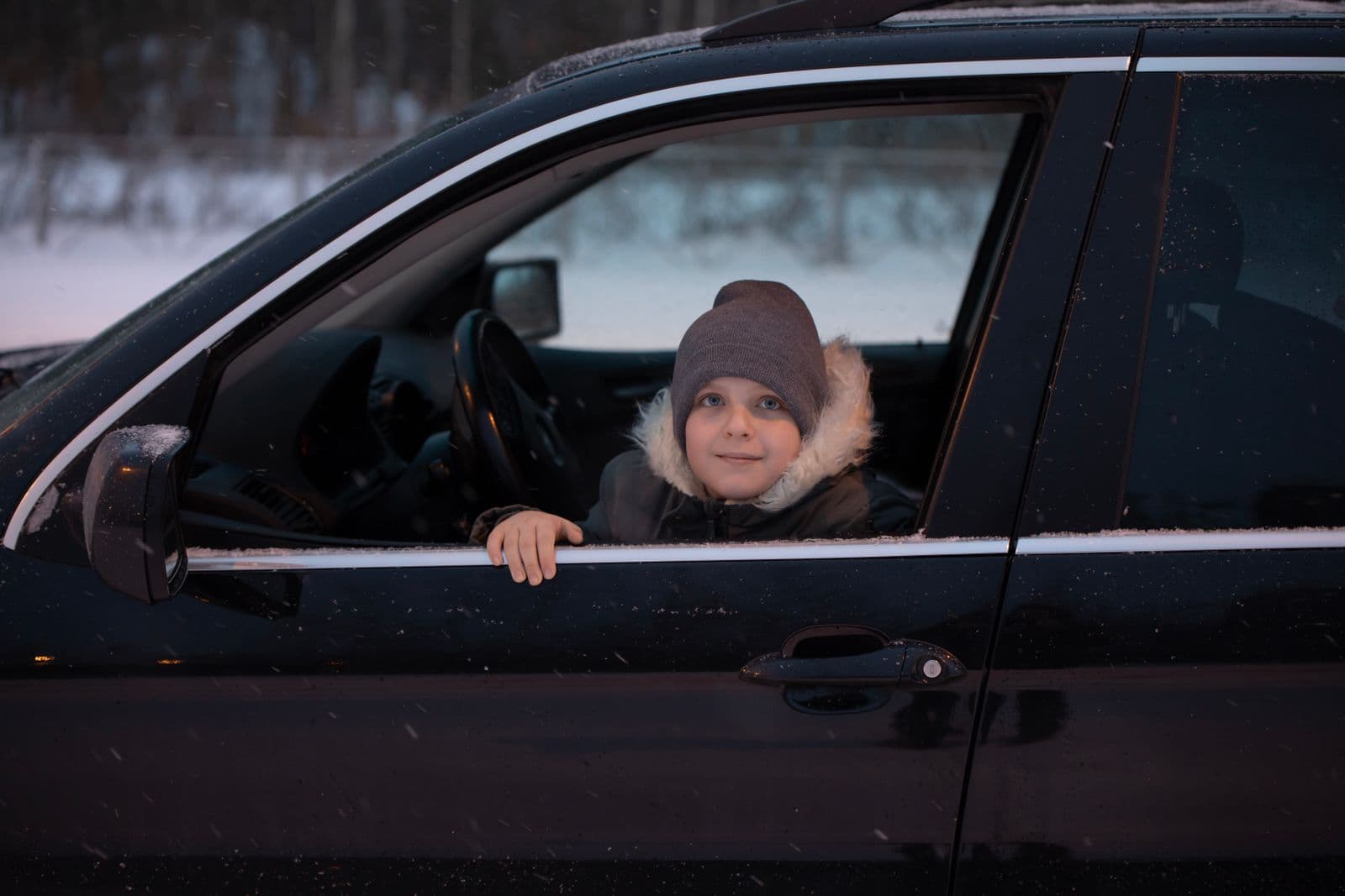 A child in a heavy hooded winter coat sitting in a car as snow falls outside