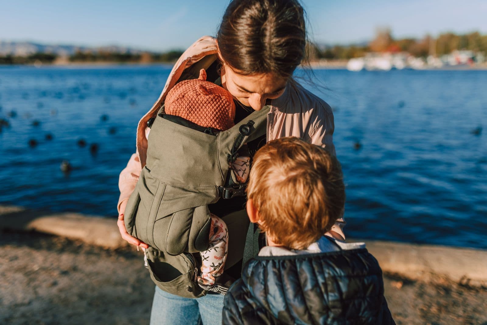 A parent carrying a baby in a structured carrier beside water with an older child nearby
